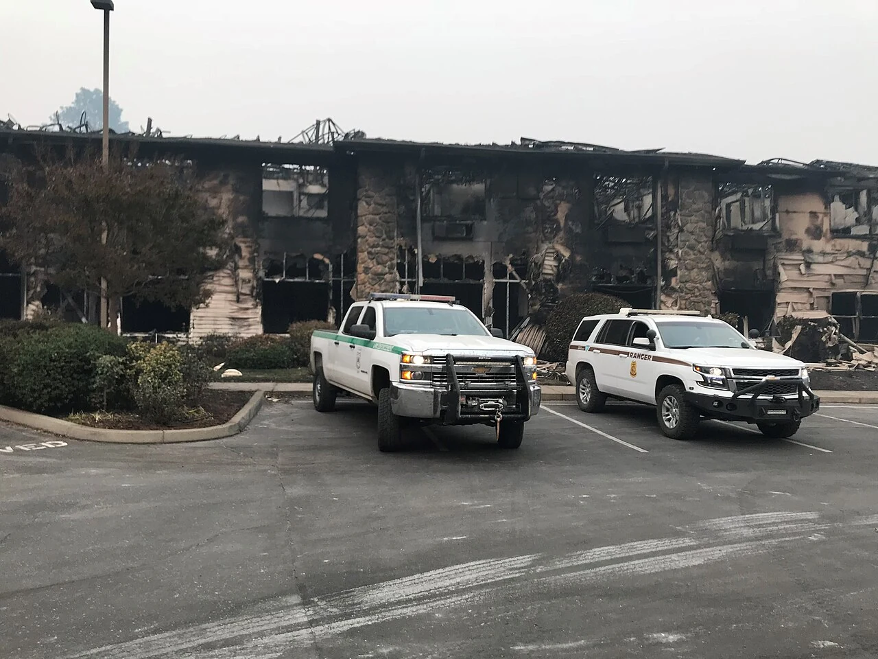 A burned-out commercial building with a collapsed roof and stone pillars still standing after the 2018 Camp Fire in Butte County, with two Forest Service vehicles parked in the empty lot
