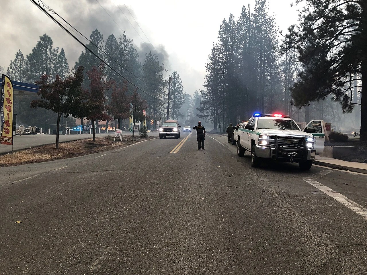 A four-lane road through Paradise, California during the 2018 Camp Fire, with smoke overhead, burning structures beside the roadway and emergency vehicles staged along the shoulder
