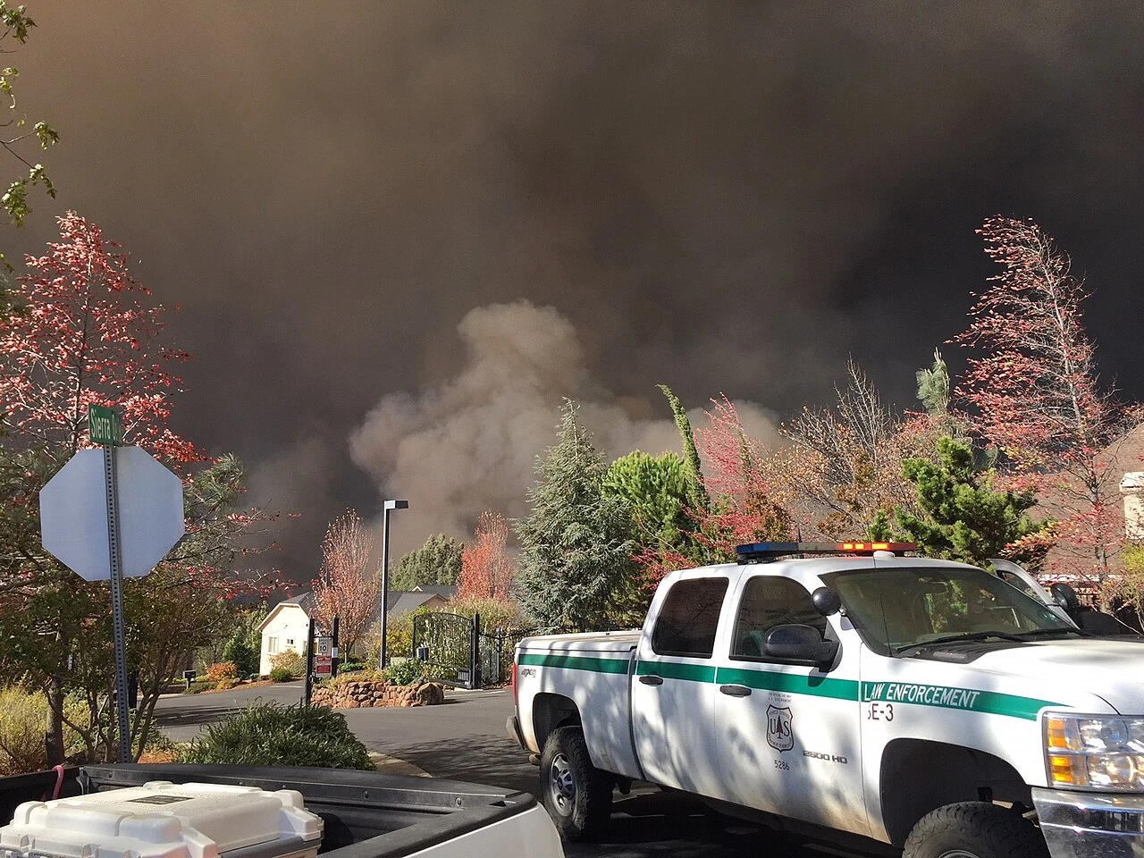 A wall of dark smoke from the 2018 Camp Fire rising directly behind houses and autumn trees on a residential street in Butte County, with a US Forest Service law enforcement truck in the foreground