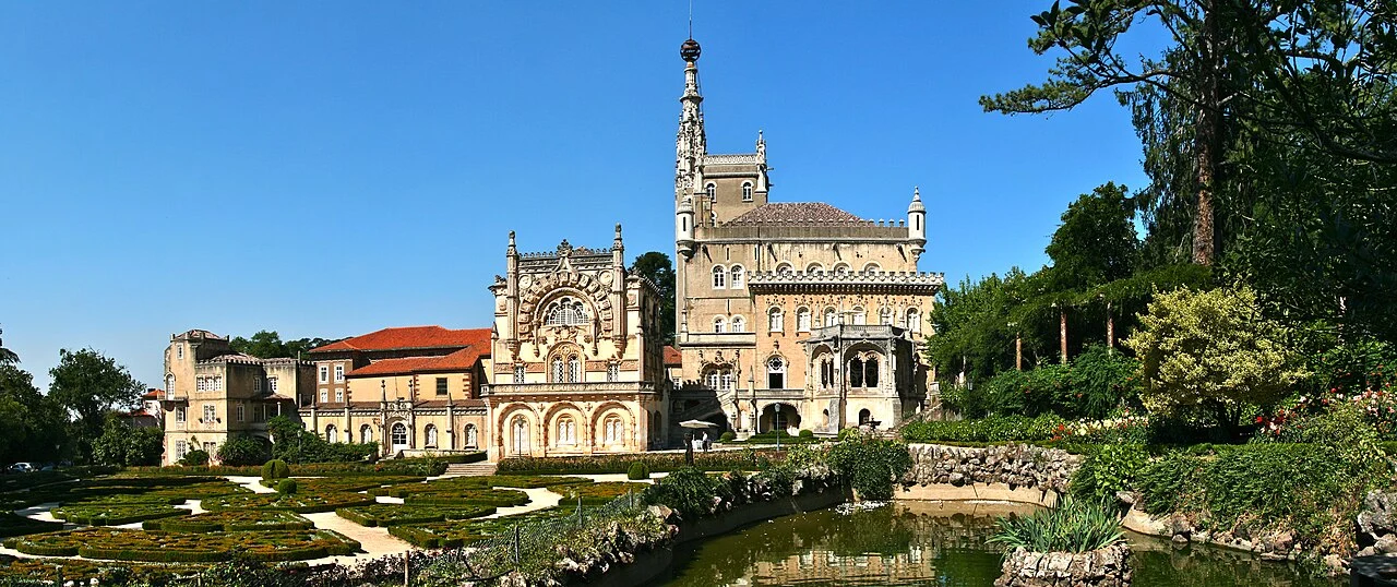 The Palace Hotel do Buçaco today: the neo-Manueline palace and its tower behind formal box-hedge gardens and a pond, with tall forest trees on either side