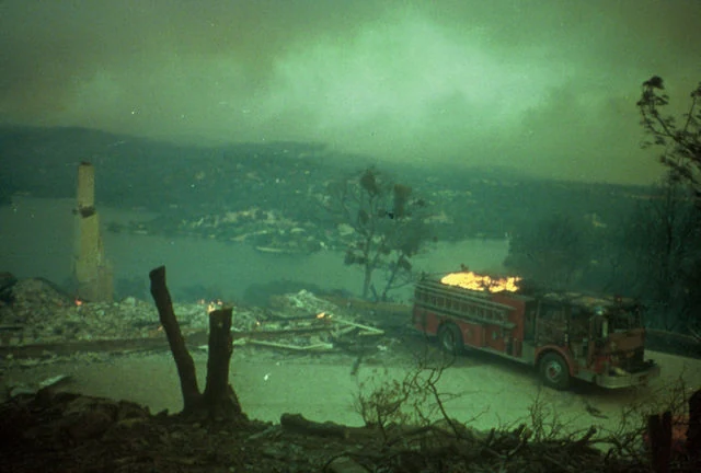 A destroyed home reduced to a chimney and rubble above a lake after the 1988 49er Fire in Nevada County, California, with a fire engine on the road beside the ruins