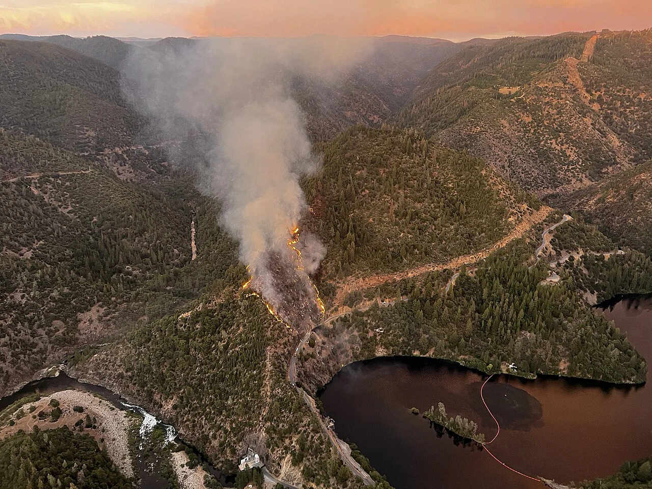 Aerial photo of the Mosquito Fire burning in steep terrain above Oxbow Reservoir near Foresthill, Placer County, on the evening of September 6, 2022, with flames and smoke rising from a forested ridge next to the reservoir