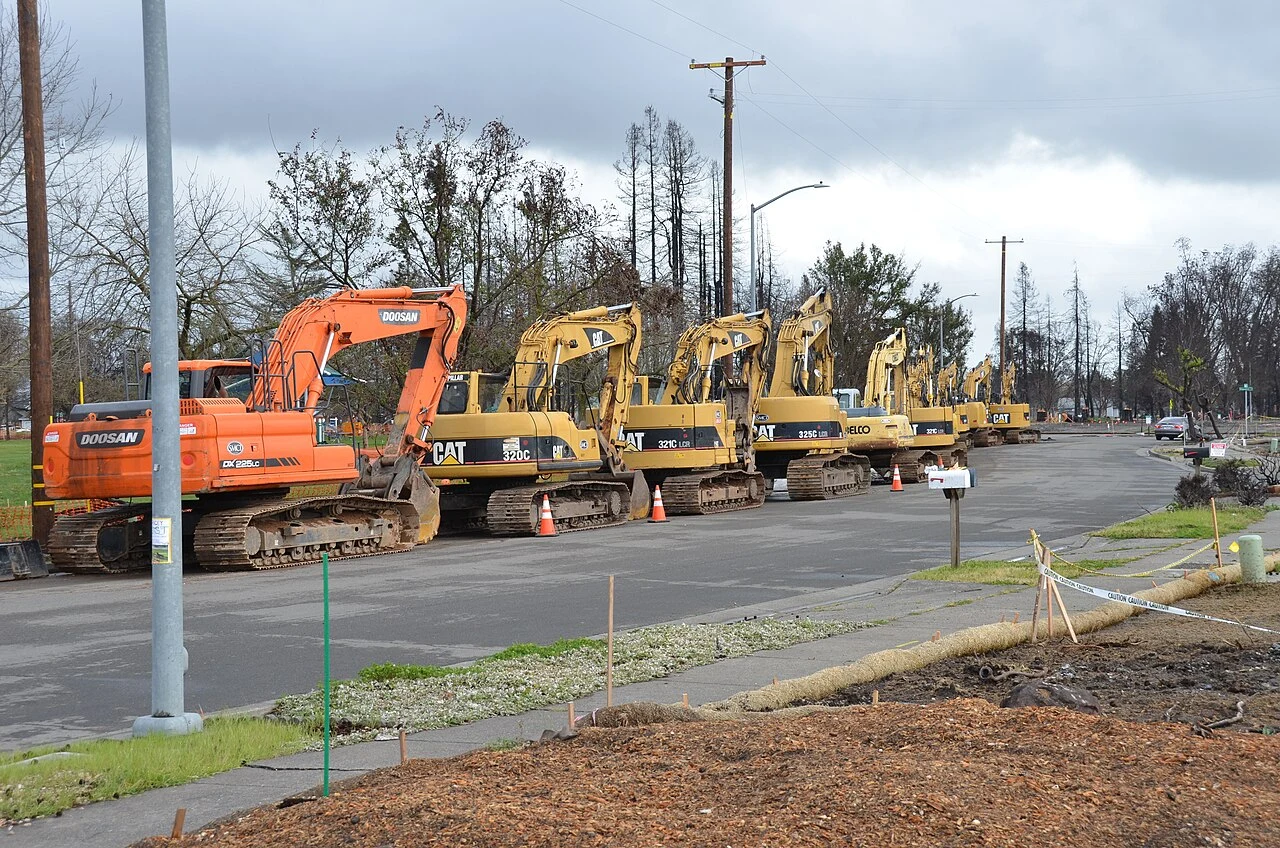 Row of excavators staged for debris removal along a burned residential street in Coffey Park, Santa Rosa, with scorched trees and cleared lots, one of the neighborhoods hardest hit by the 2017 Tubbs Fire